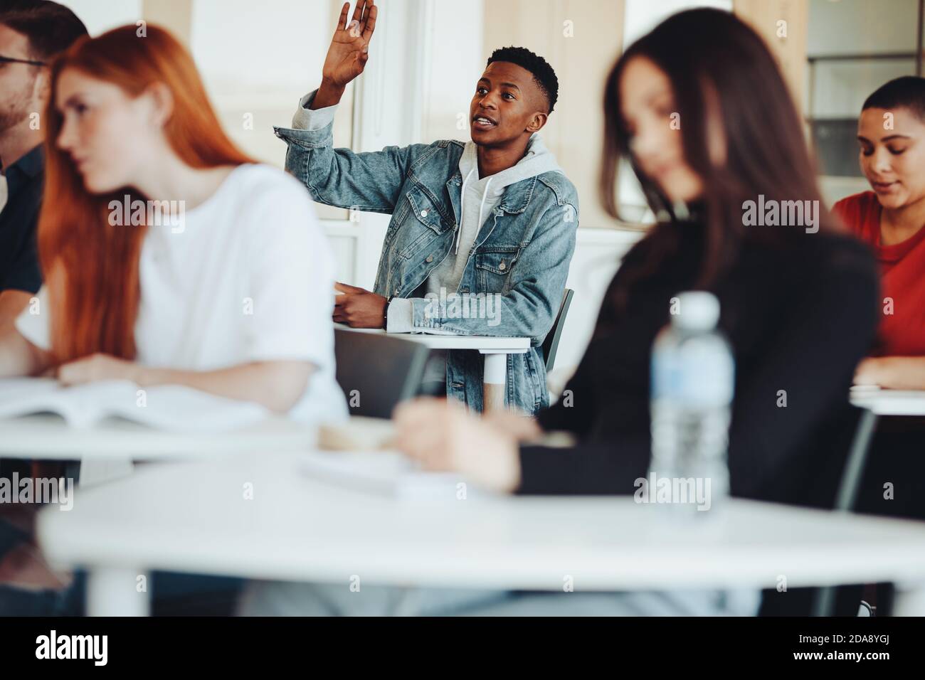 Studente maschile seduto in classe e alzando la mano per fare una domanda durante la lezione. Studente universitario che chiede una domanda al docente in classe. Foto Stock