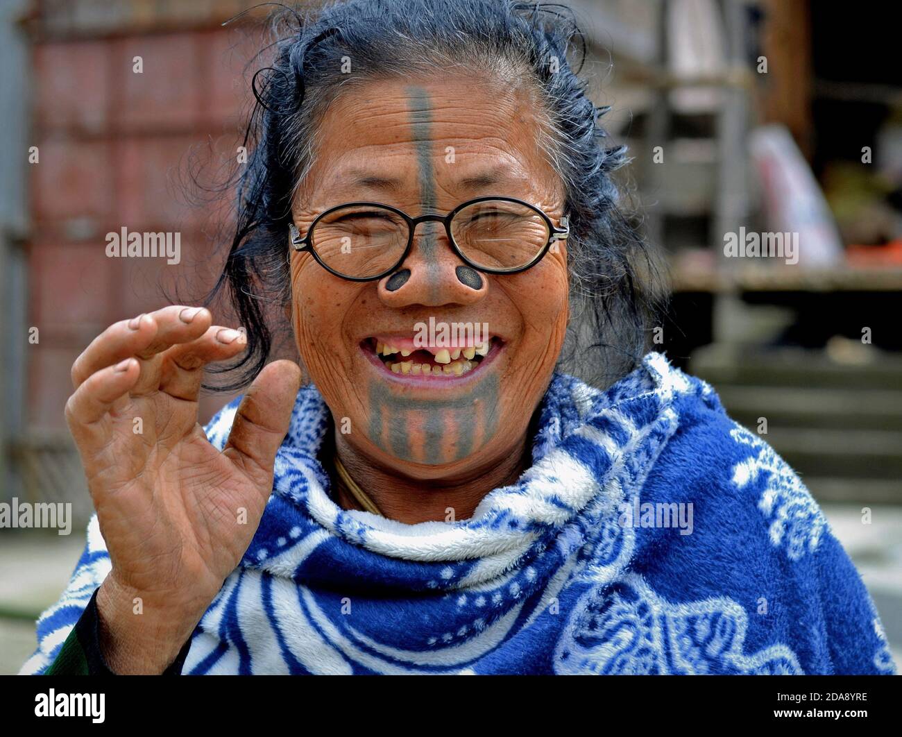 La vecchia tribale dell'India nordorientale Apatani con i tappi neri del naso di legno e il tatuaggio tradizionale del viso indossa gli occhiali moderni e sorrisi per la macchina fotografica. Foto Stock