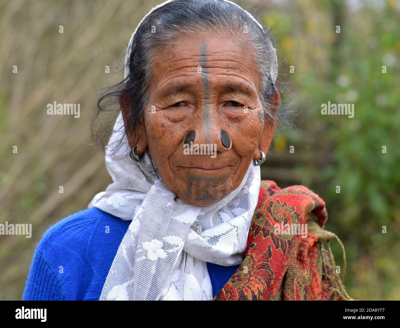 Vecchia donna tribale di minoranza etnica Apatani dell'India nordorientale con i tappi neri del naso di legno e i tatuaggi tradizionali del viso pone per la macchina fotografica. Foto Stock