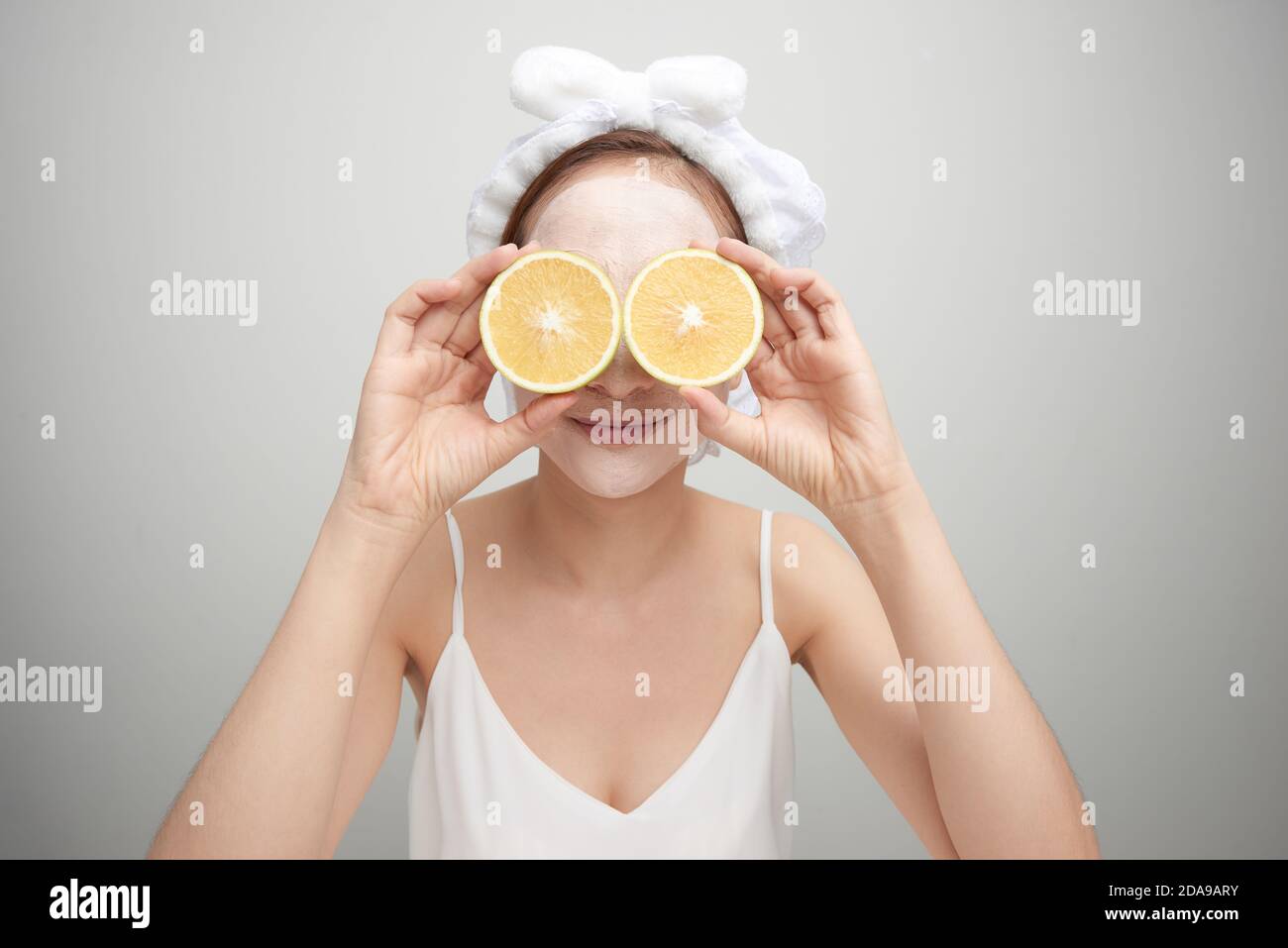 Giovane donna con maschera facciale in argilla che tiene una fetta di frutta arancione che copre gli occhi Foto Stock
