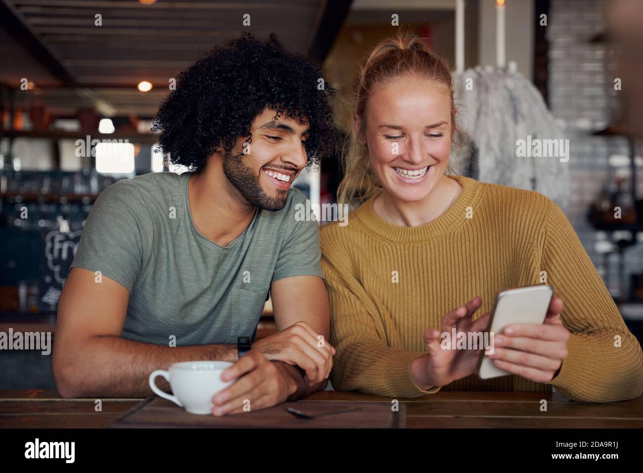 Giovane donna allegra che mostra video divertente in smartphone al ragazzo mentre si rilassa e si siede in un moderno cafe' Foto Stock
