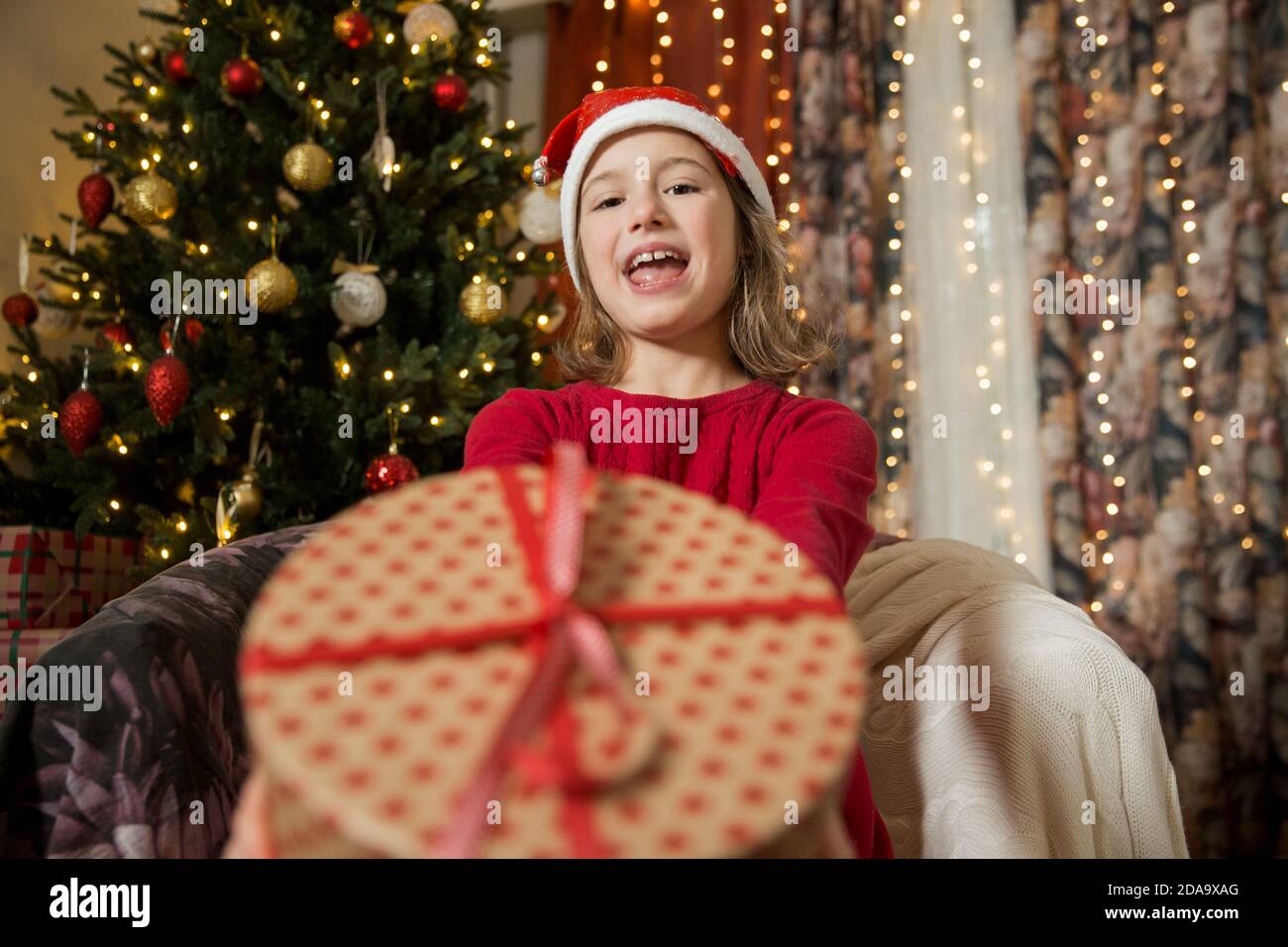 Un bambino felice nel cappello rosso di Santa sta celebrando il Natale. Carina ragazza che tiene avvolto dono, sorridente e ondeggiante mano, seduta in decorato con Natale Foto Stock