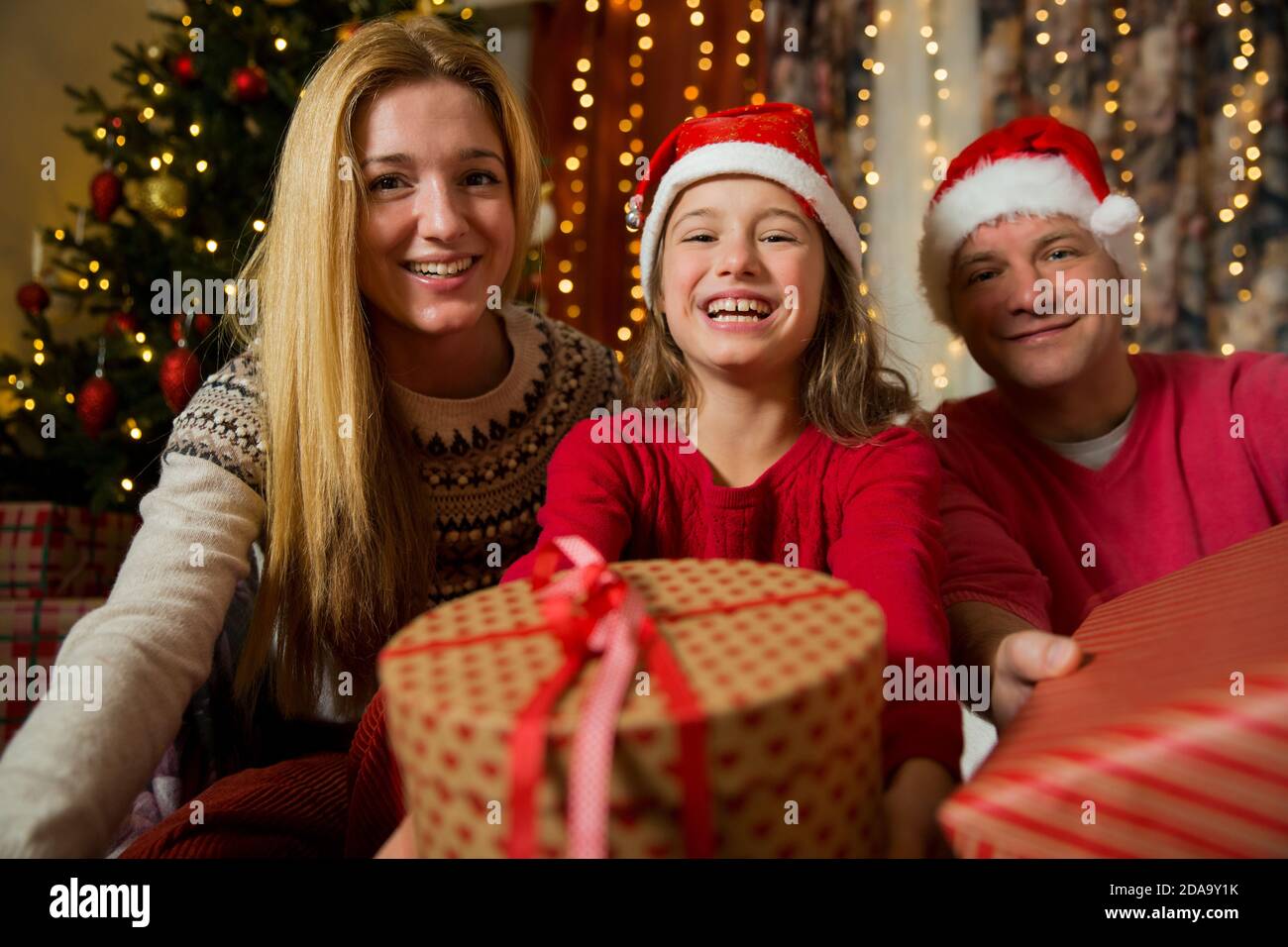 Una famiglia felice con il bambino nel cappello rosso di Santa sta celebrando il Natale. Ragazza carina che tiene il regalo avvolto, sorridente e ondeggiante mano Foto Stock