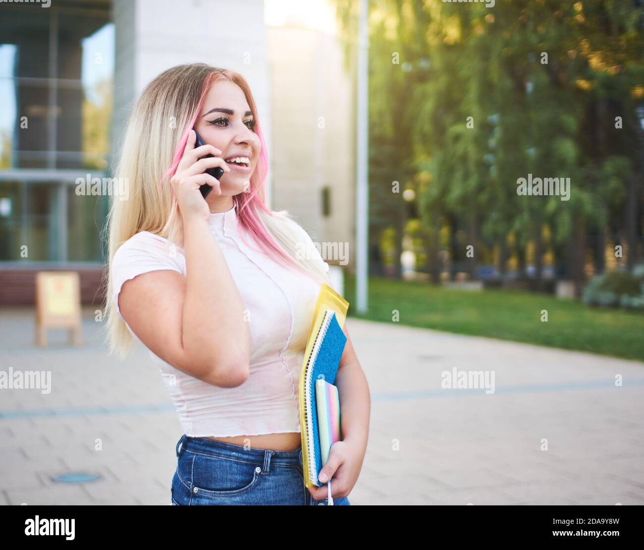 Sorridente giovane studentessa di generazione z in piedi in un campus di fronte alla facciata di un edificio universitario e che parla sullo smartphone Foto Stock