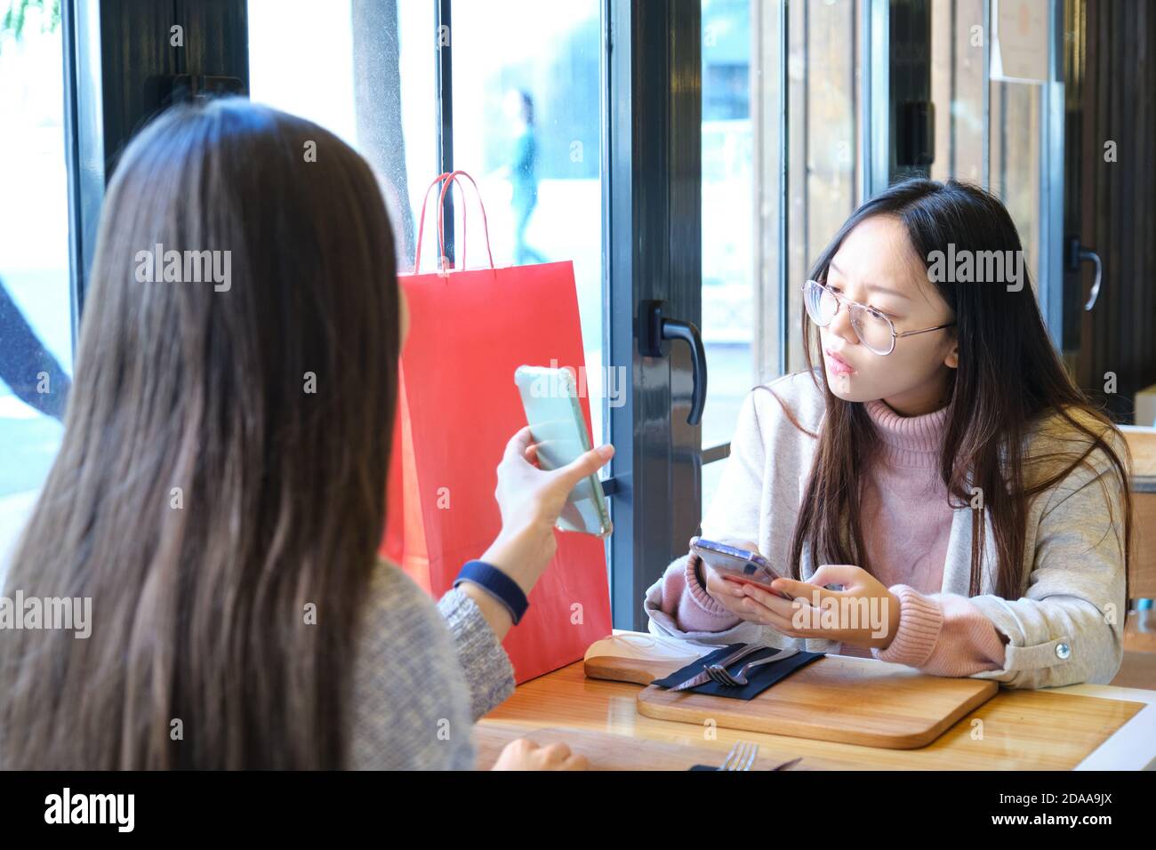 Giovane donna caucasica che mostra qualcosa al suo amico asiatico nel suo smartphone dopo lo shopping. Donne caucasiche e asiatiche in un ristorante. Foto Stock