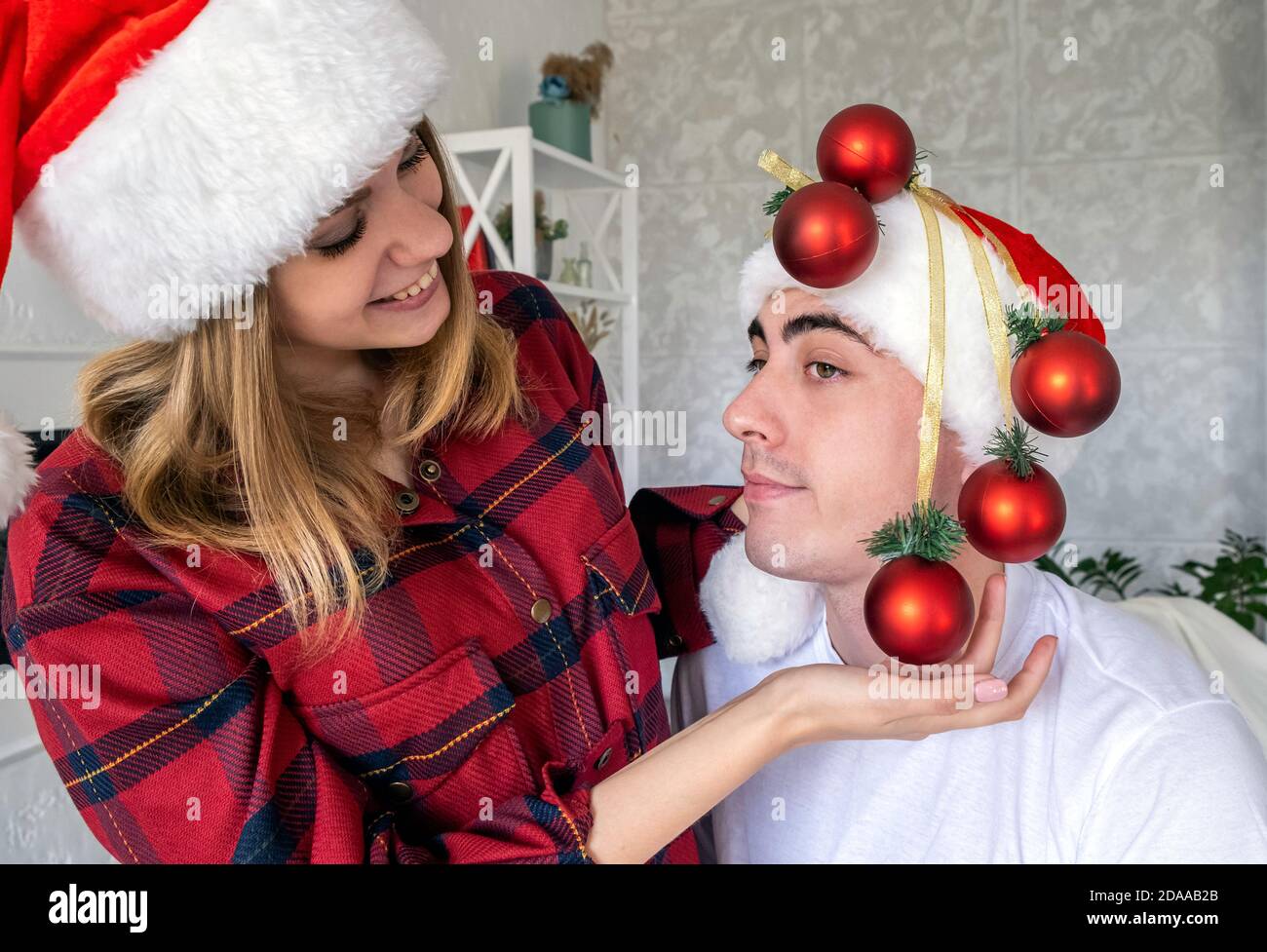 Giovane giocosa donna che si fooling intorno a decorare l'uomo con le palle di Natale invece dell'albero di Natale. Coppia che indossa cappelli santa e pigiami rossi. Foto Stock