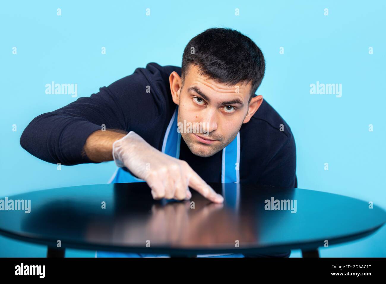 Un uomo in guanti di gomma sfrega con il dito e controlla la pulizia del tavolo. Un uomo in un grembiule controlla il lavoro domestico. Stereotipo di genere. Foto Stock