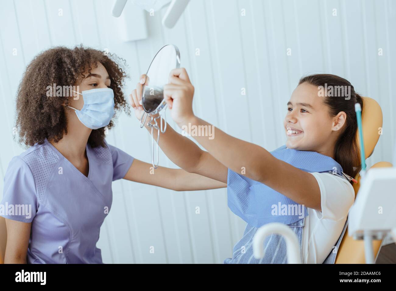 Felice piccola ragazza guarda nello specchio per stimare il lavoro del dentista e godendo il risultato Foto Stock