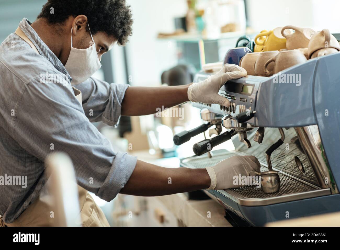 Millenario afroamericano uomo in grembiule, guanti e maschera protettiva funziona con macchina da caffè al mattino in interni Foto Stock