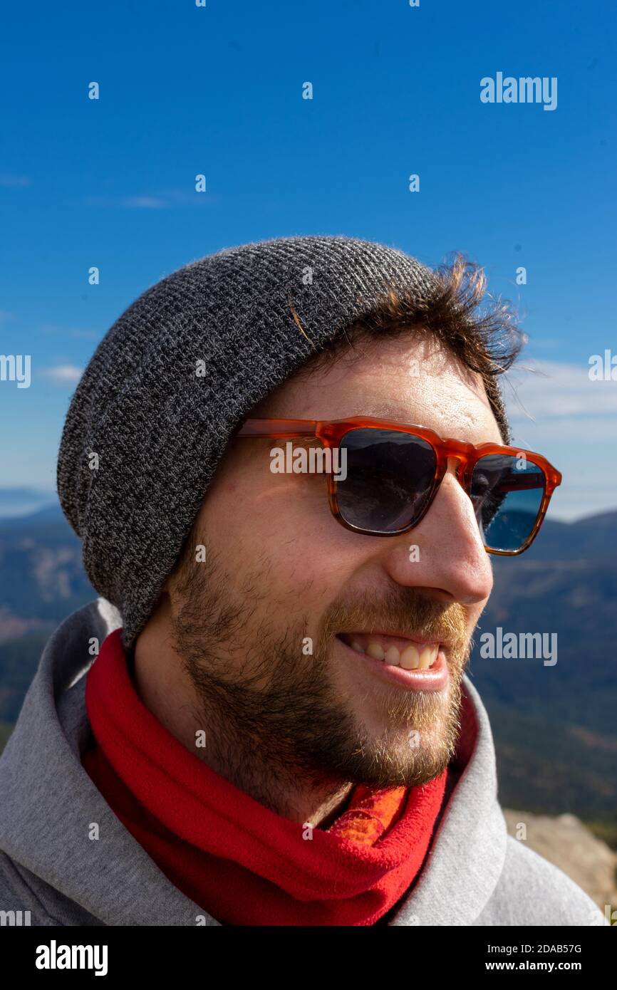 Attraente giovane uomo bianco con una barba, occhiali da sole e uno stile casual sorride all'aperto in un ambiente naturale soleggiato Foto Stock