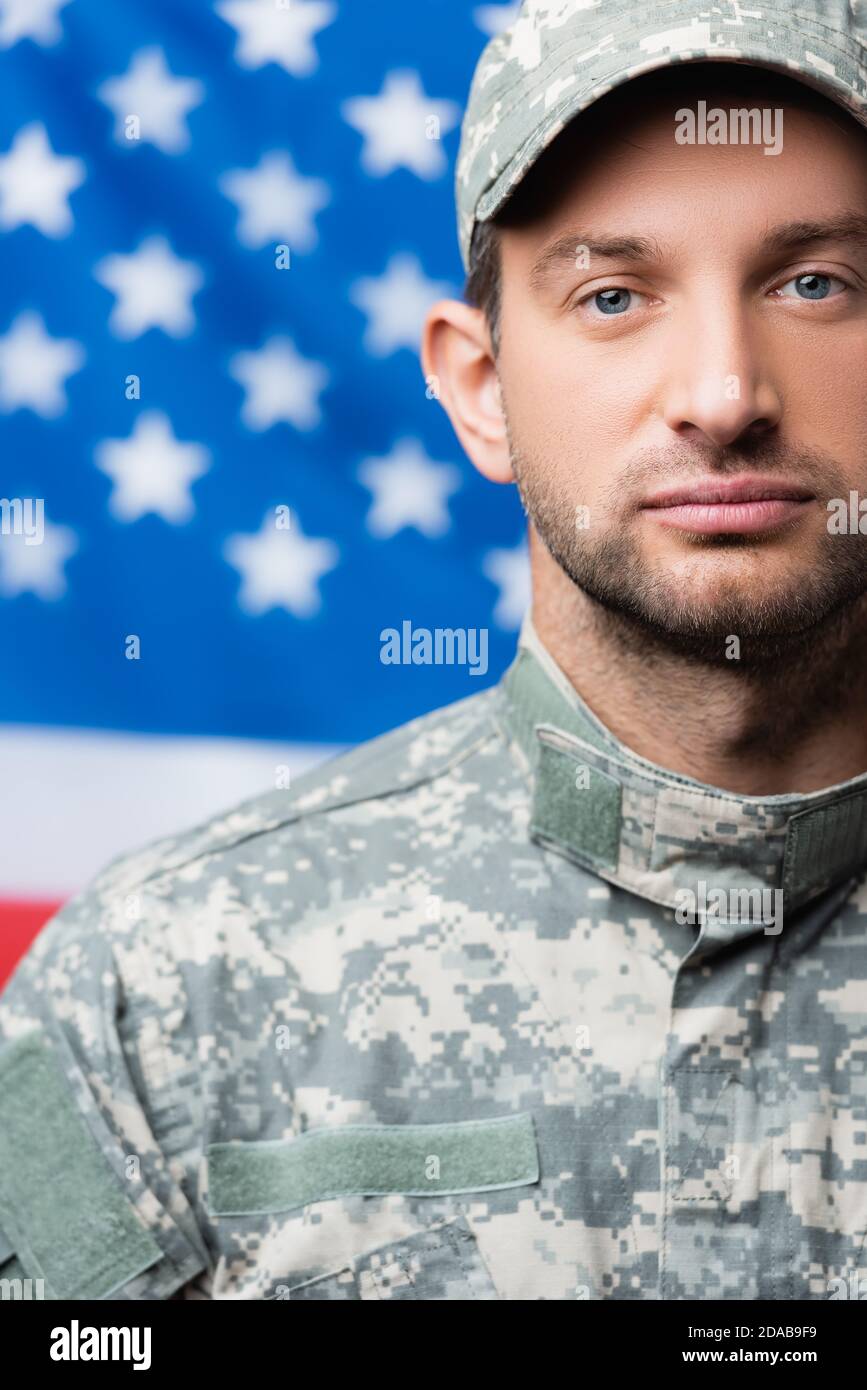 uomo militare in uniforme guardando la macchina fotografica vicino alla bandiera americana su sfondo sfocato Foto Stock