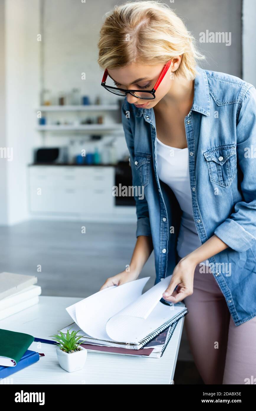 giovane donna bionda in occhiali che lavorano da casa e girando pagine del notebook Foto Stock