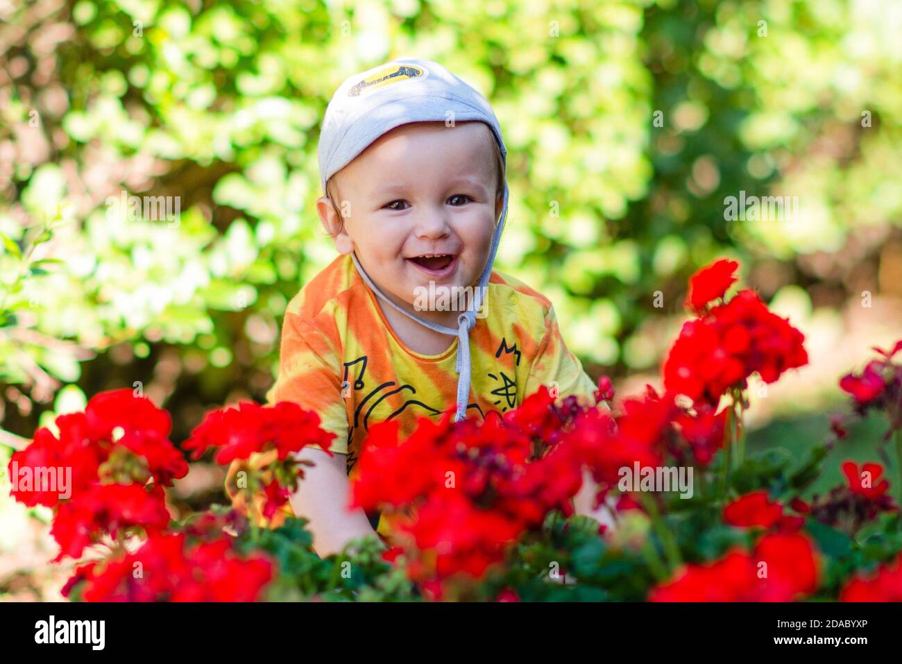 Felice bambino sorridente con cappello sorrondato da fiori rossi e. siepe verde Foto Stock