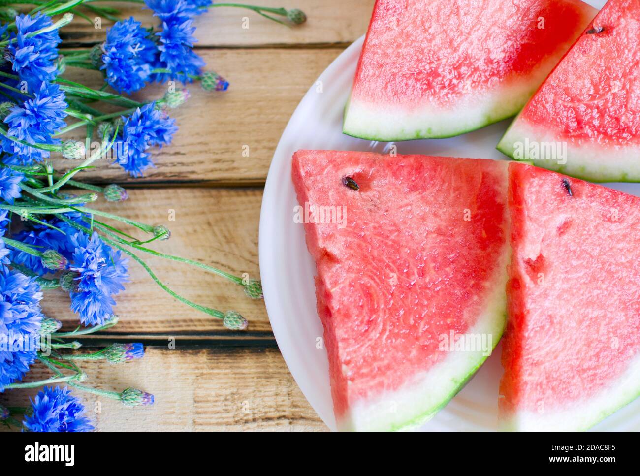 Fiori estivi di stagione (fiori di mais blu) e frutta (cocomero e fragole) su tavolo rustico in legno scottato Foto Stock