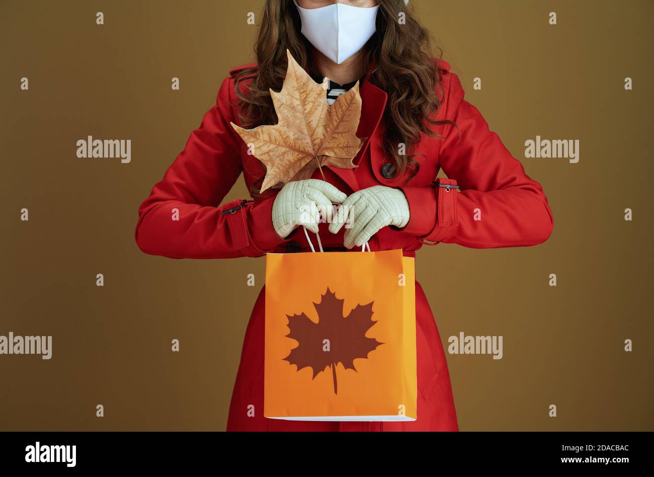 Vita durante la pandemia del coronavirus. Primo piano su donna di mezza età in camice rosso con foglia di acero giallo autunno e borsa di carta isolato sul dorso marrone Foto Stock