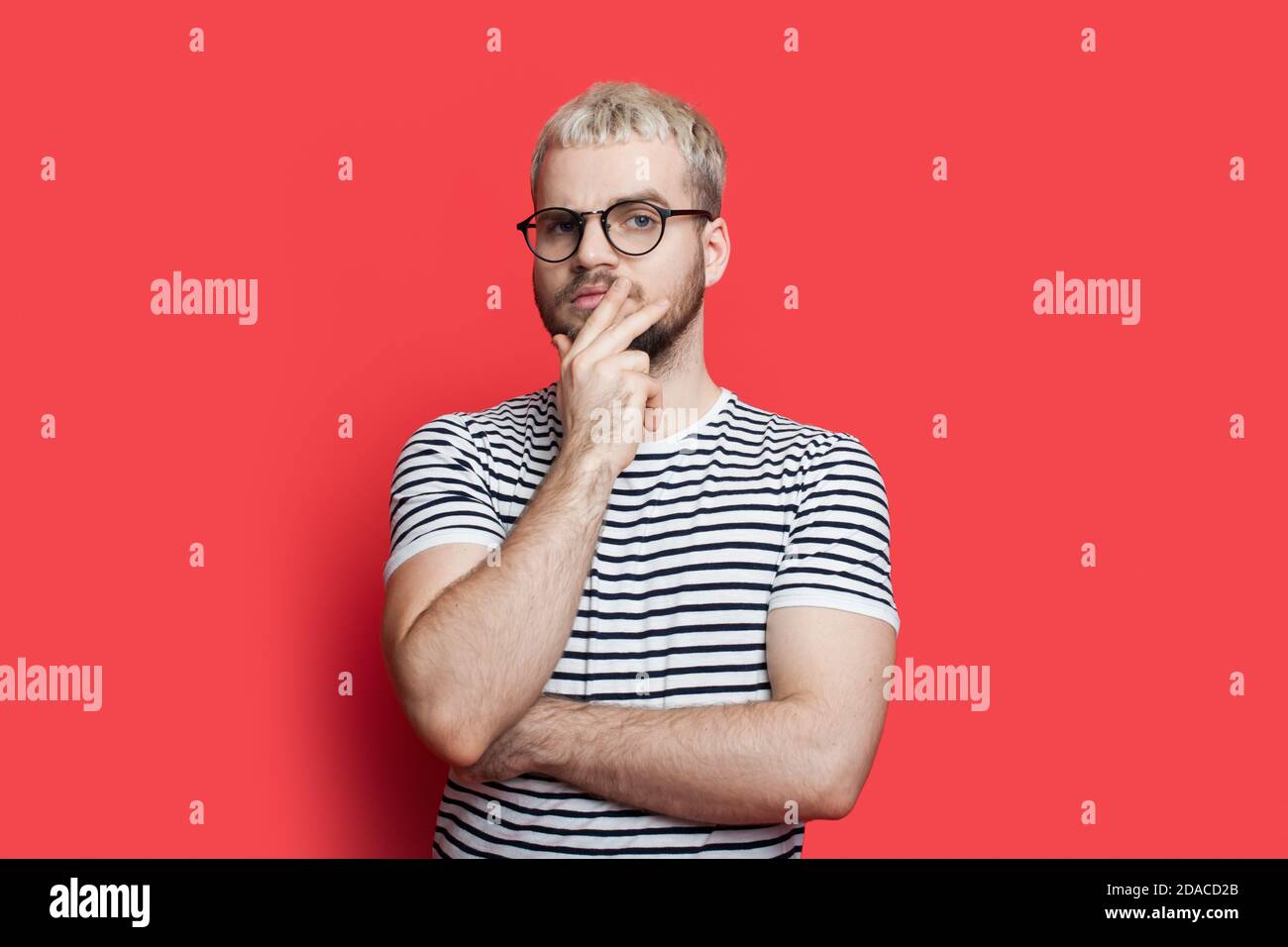 L'uomo caucasico biondo con barba e occhiali è in posa premuroso toccando il mento su una parete rossa dello studio guardando fotocamera Foto Stock