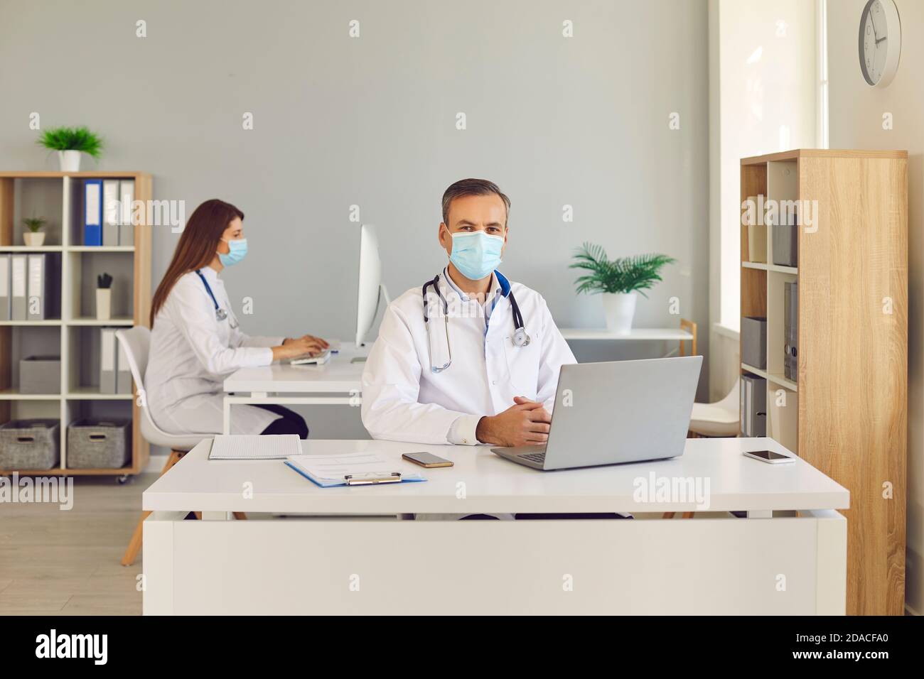 Medico maschile in maschera medica protettiva seduto sul posto di lavoro con il computer in ufficio. Foto Stock