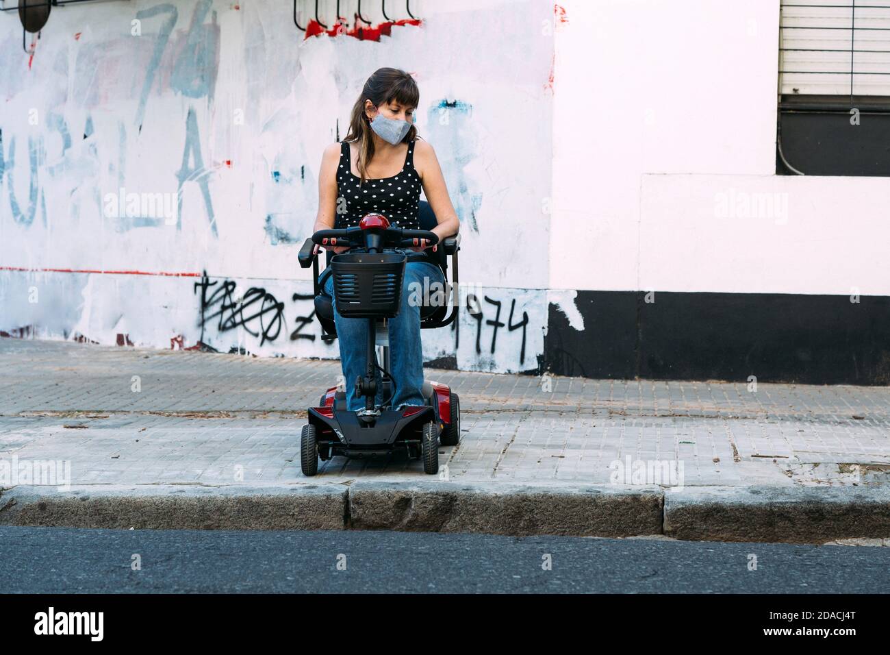 Giovane donna in scooter elettrico per disabili. Non può attraversare la strada perché non c'è rampa. Grunge parete sullo sfondo. Invaso Foto Stock