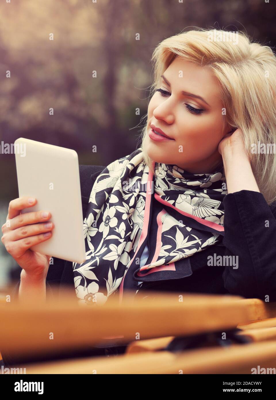 Giovane biondo business donna con tavoletta digitale computer seduta sul banco di moda elegante modello femminile indossare nero classico cappotto e sciarpa Foto Stock