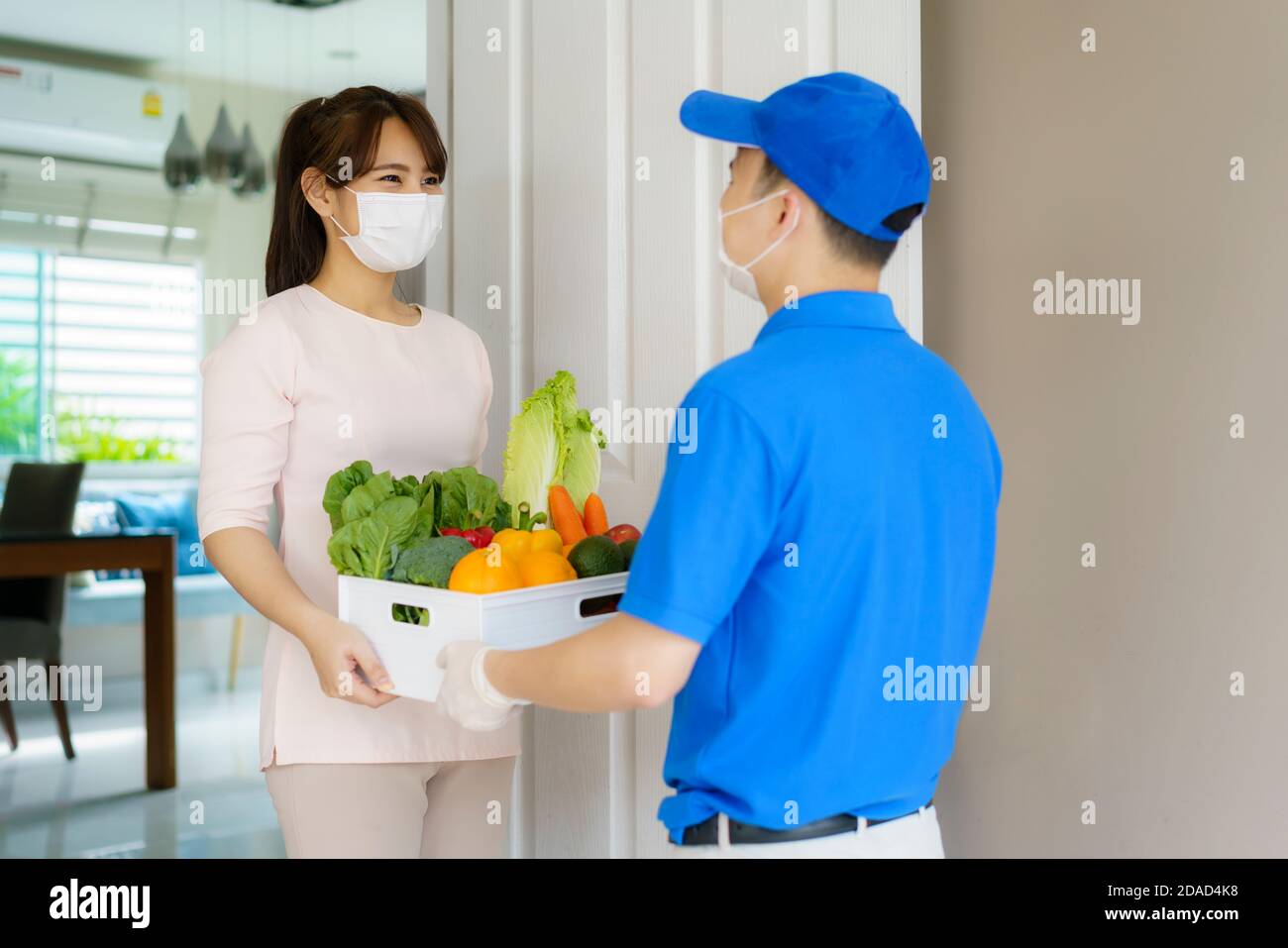 Donna asiatica costumer indossare maschera viso ricevere alimentari scatola di cibo, frutta e verdura da uomo di consegna di fronte alla casa durante il periodo di hom Foto Stock