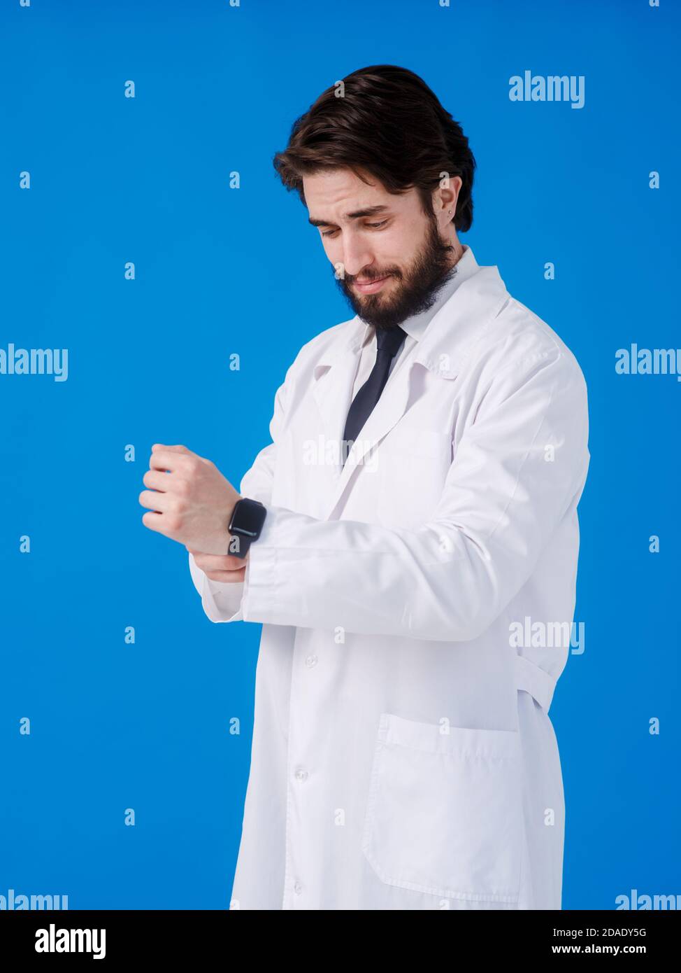 Bell'uomo medico con barba indossata e stetoscopio su sfondo blu controllare il tempo sull'orologio da polso, rilassato e sicuro Foto Stock