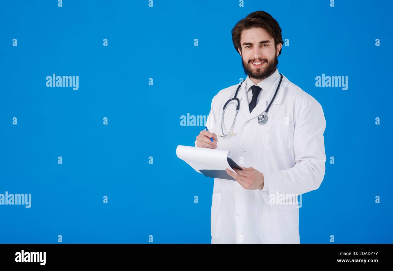 un giovane medico con la bearded in un cappotto medico bianco sopra uno sfondo blu scrive i reclami del paziente su un tablet con documenti e medici Foto Stock