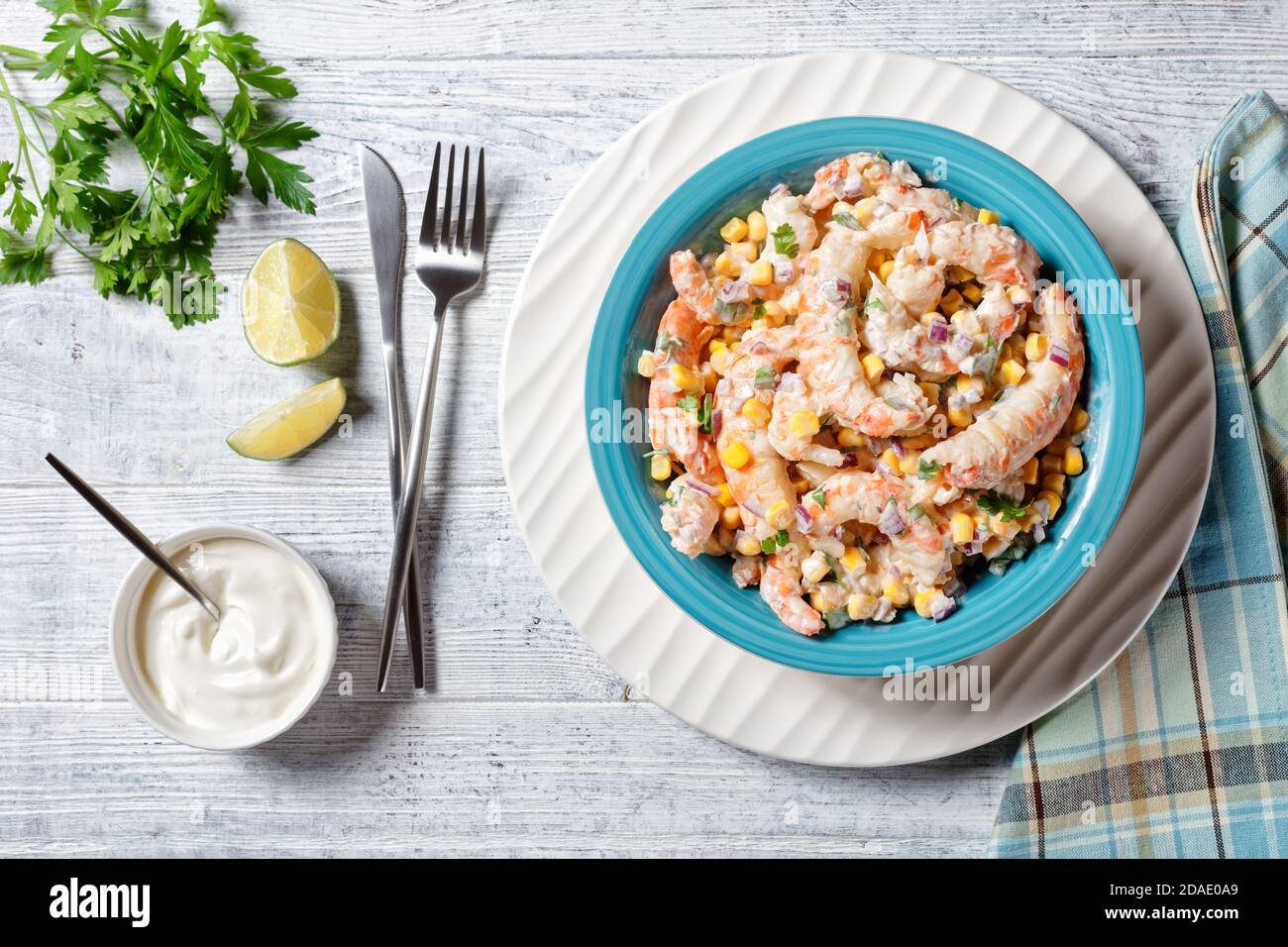 Insalata di gamberi con salsa di panna acida e succo di lime, mais, cipolla rossa e prezzemolo serviti su un recipiente per insalata blu su un piatto di ricarica bianco con posate o Foto Stock