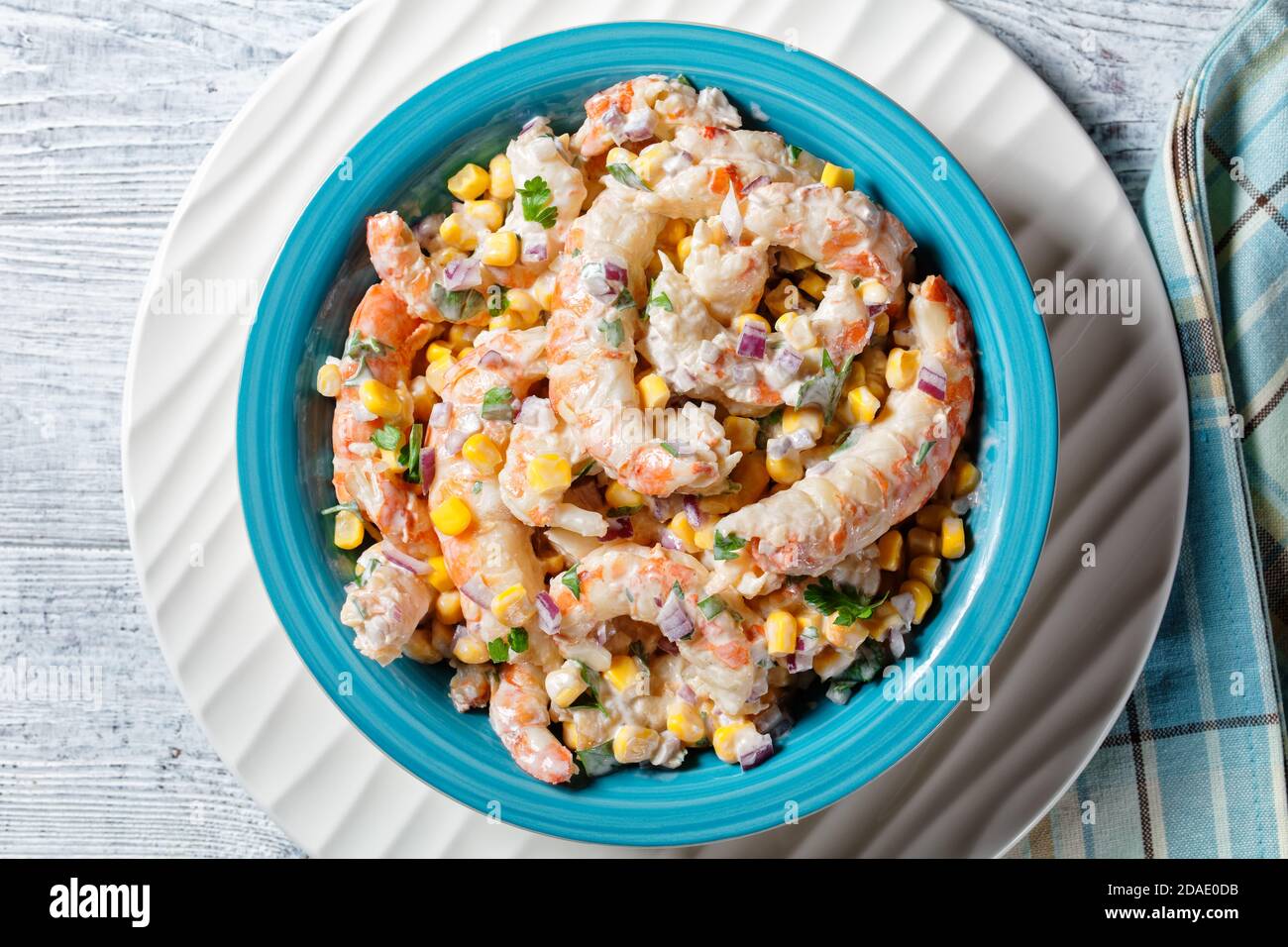 Insalata di gamberi con salsa di panna acida e succo di lime, mais, cipolla rossa e prezzemolo serviti su un recipiente per insalata blu su un piatto di ricarica bianco con posate o Foto Stock