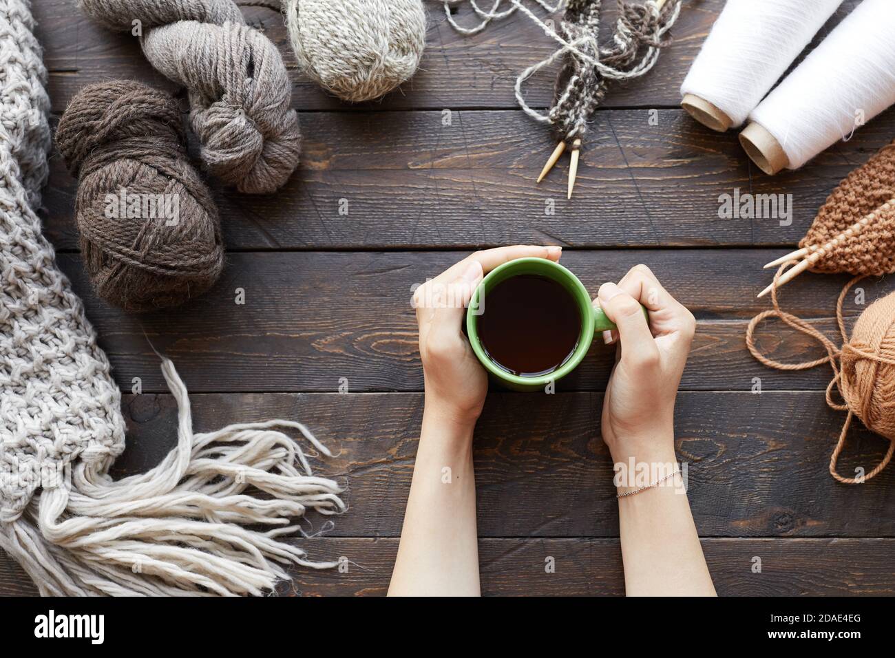 Primo piano di donna seduta al tavolo di legno con palle di filati e tessuti di lana finiti e tè caldo da bere Foto Stock