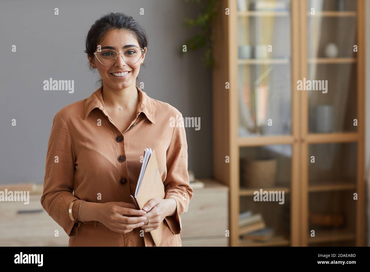 Ritratto di giovane donna d'affari in occhiali e in blusa in presa cartelle e sorridendo alla fotocamera in ufficio Foto Stock
