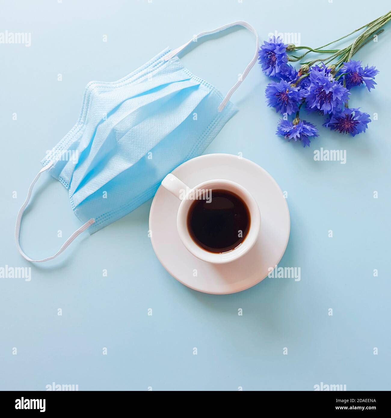 Tazza di caffè, fiori blu e blu rimosso maschera medica chirurgica protettiva. Colazione nel caffè. Realtà moderna e concetto di vita. Vista dall'alto, piatta Foto Stock