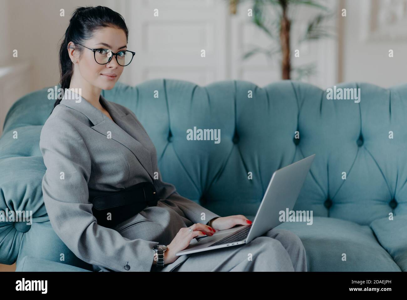 Financer femminile di successo aggiorna i dati in applicazione sul computer portatile moderno, guarda seriamente alla fotocamera, si siede su comodo divano, possiede il progetto di Foto Stock