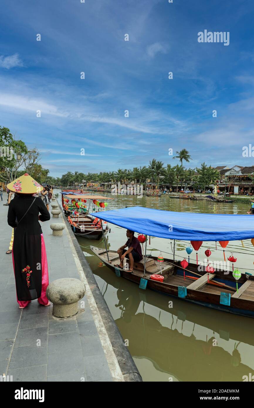 Una donna con un tradizionale cappello conico vietnamita, una non la, in piedi su Bach Dang Street accanto a una barca sul fiume Thu Bon, Hoi An, Vietnam, Asia Foto Stock