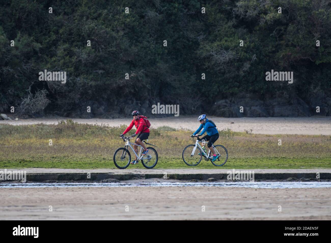 Due ciclisti che cavalcano le loro biciclette lungo il fiume esposto del fiume Gannel a bassa marea a Newquay in Cornovaglia. Foto Stock