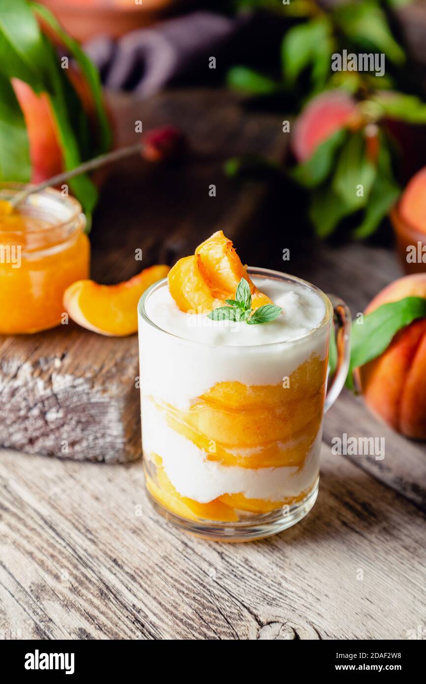 Dessert alla frutta di pesca in una tazza di vetro su un tavolo rustico in legno con frutta fresca di pesca, marmellata di pesche. Dessert fatto in casa con frutta. Macedonia di frutta con yogurt o. Foto Stock