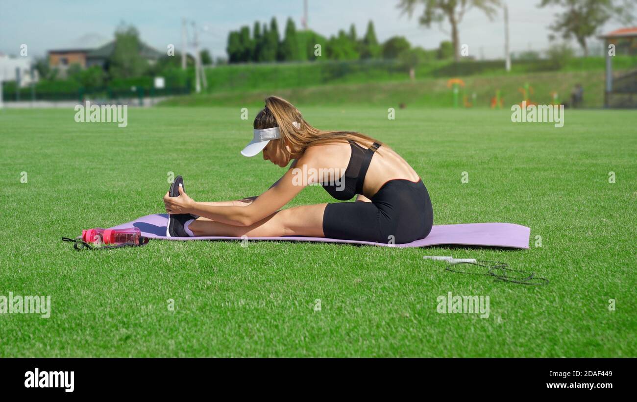 Vista laterale della giovane donna che allunga le gambe sul tappetino dopo la corsa allo stadio nella giornata di sole d'estate. Ragazza attraente che indossa vestito nero facendo esercizi rilassanti all'aperto. Flessibilità, concetto di allenamento. Foto Stock