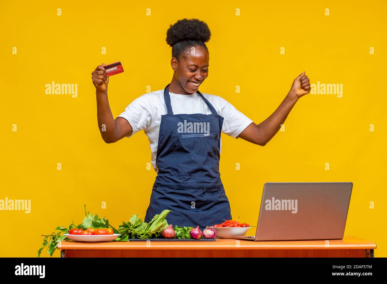 lo chef africano eccitato gioisce nel bel mezzo della preparazione di un pasto tenendo la sua carta di credito e utilizzando il suo computer portatile computer Foto Stock