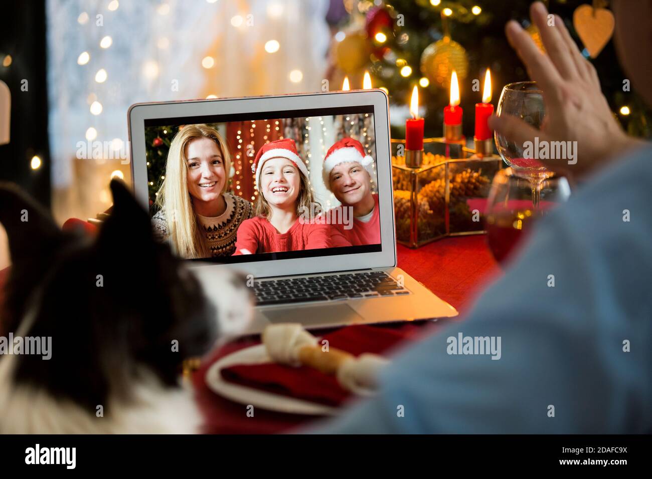 Uomo maturo che festeggia il Natale con il suo cane seduto al tavolo servito con il computer portatile. Le persone salutano i loro amici durante una videochiamata utilizzando la webcam. Foto Stock