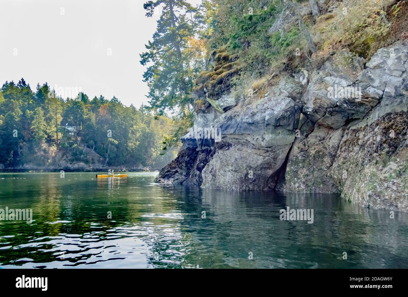In lontananza, un kayak soleggiato si trova in piena luce del sole, a differenza del primo piano, dove un promontorio roccioso e mussoso si trova all'ombra profonda (costa della British Columbia). Foto Stock