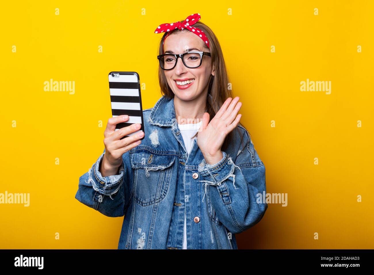 Giovane donna con giacca in denim e occhiali per un telefono nelle sue mani e parla di chat video su un sfondo giallo Foto Stock