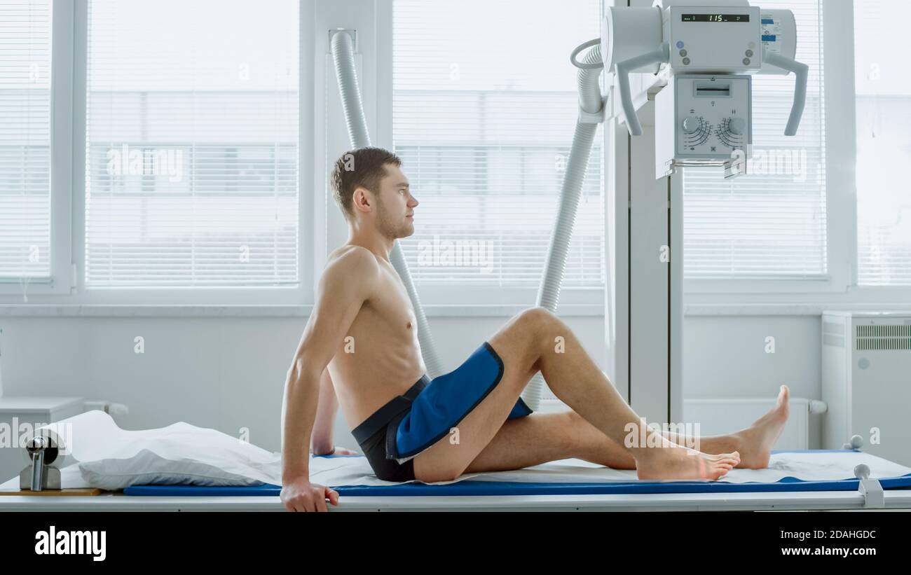 Nell'ospedale, vista laterale dell'uomo seduto su un letto in attesa che la macchina a raggi X possa eseguire la scansione della gamba per eventuali lesioni. Ricerca di fratture, arti rotti Foto Stock