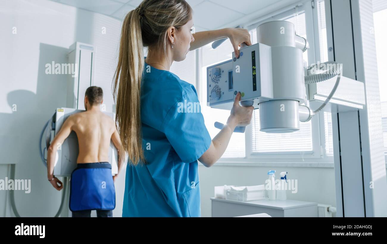 Nell'ospedale, l'uomo in piedi contro il muro, mentre il tecnico medico regola la macchina a raggi X per la scansione. Ricerca di fratture, arti rotti Foto Stock