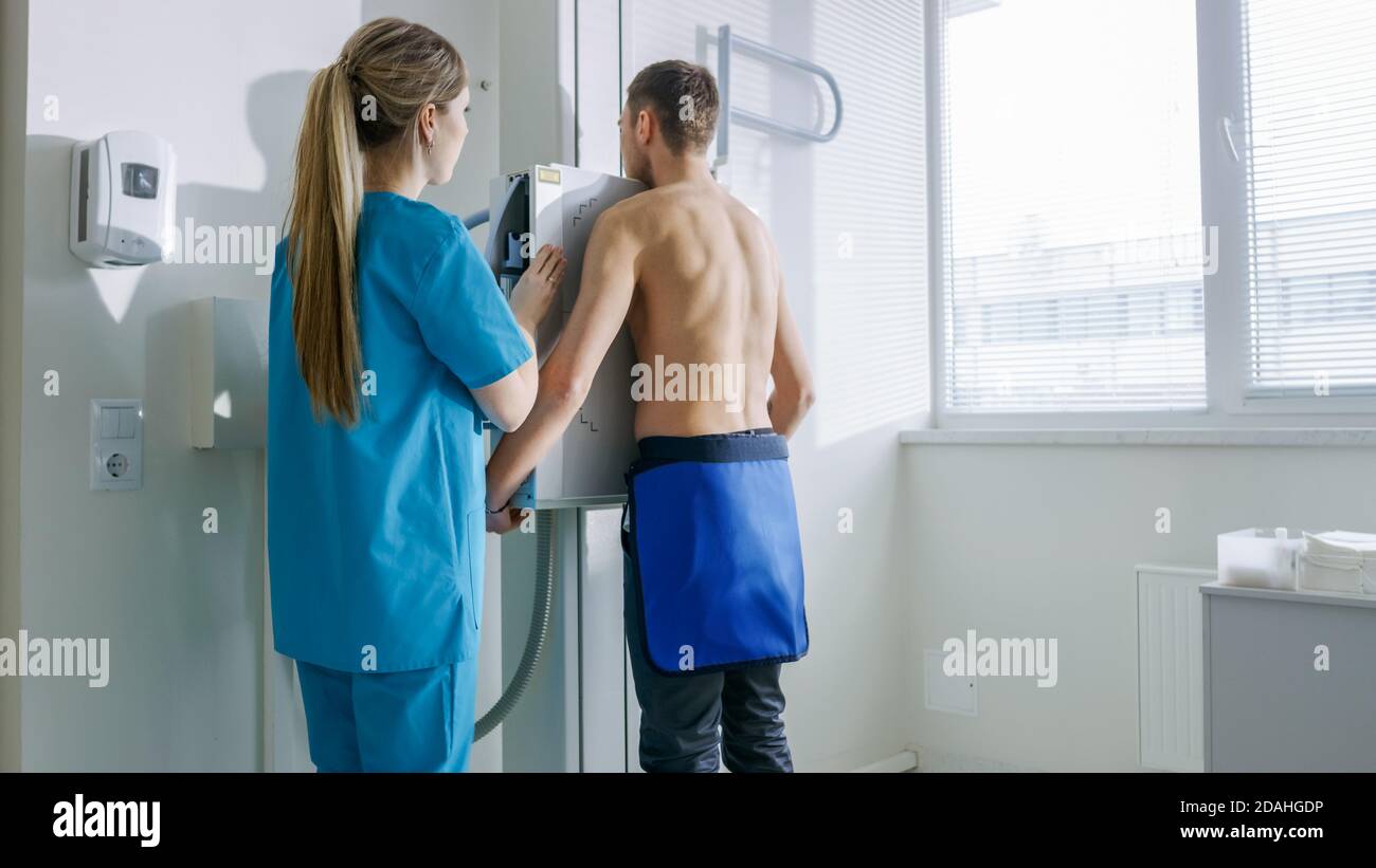 Nell'ospedale, l'uomo in piedi contro il muro, mentre il tecnico medico regola la macchina a raggi X per la scansione. Ricerca di fratture, arti rotti Foto Stock