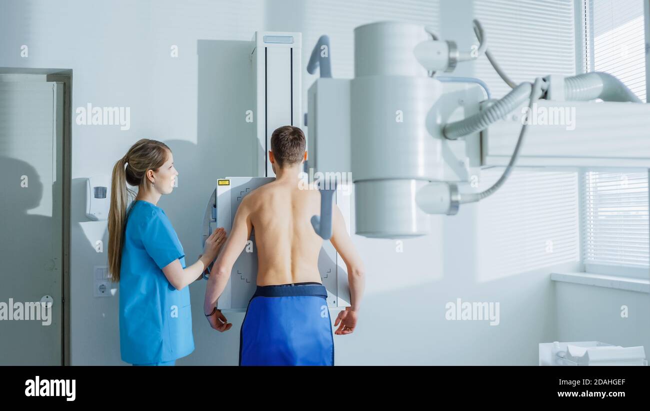 Nell'ospedale, l'uomo in piedi contro il muro, mentre il tecnico medico regola la macchina a raggi X per la scansione. Ricerca di fratture, arti rotti Foto Stock