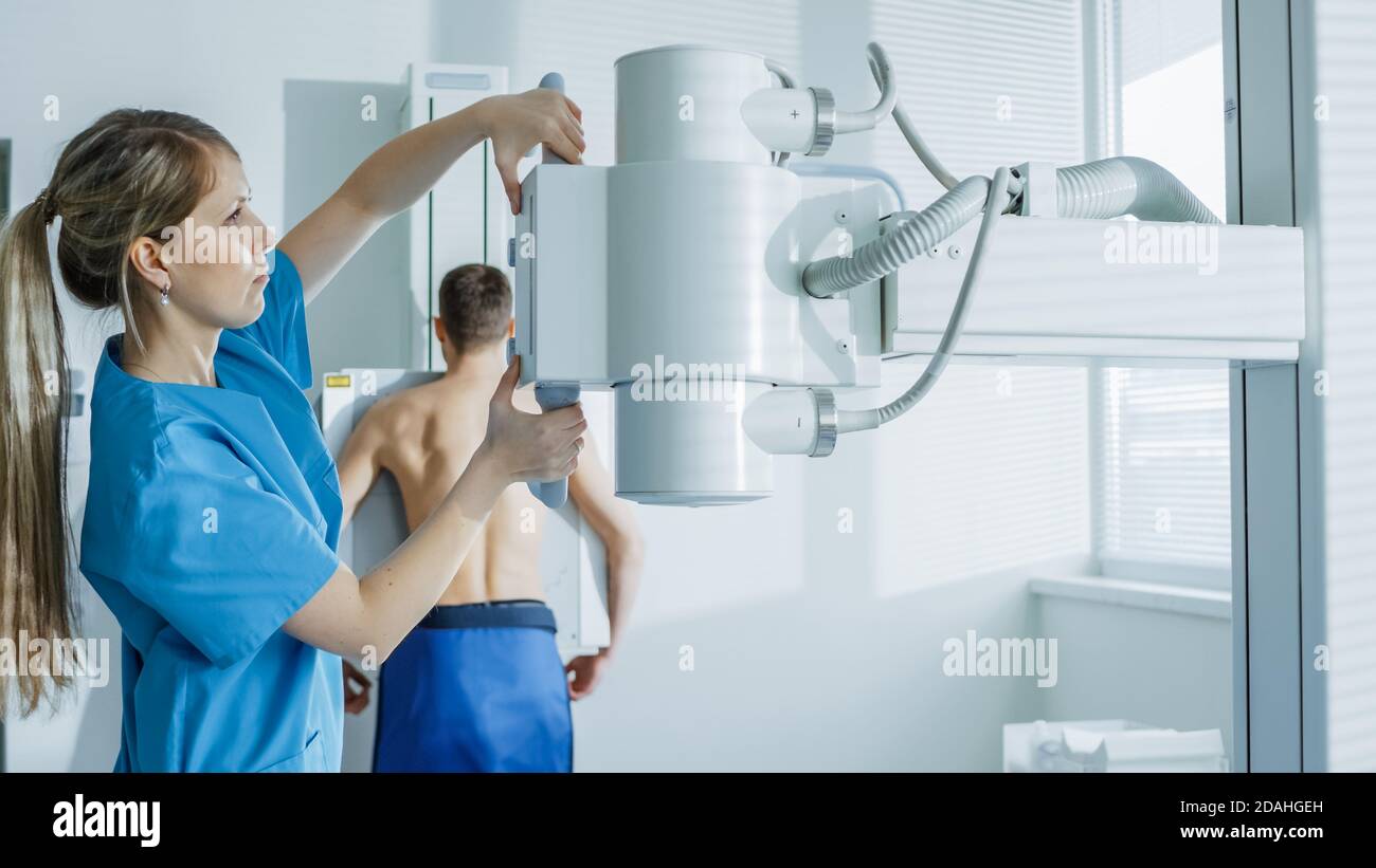 Nell'ospedale, l'uomo in piedi contro il muro, mentre il tecnico medico regola la macchina a raggi X per la scansione. Ricerca di fratture, arti rotti Foto Stock