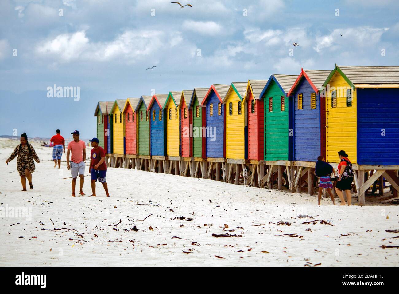 Muizenberg, Città del Capo, Sudafricana - 31 DICEMBRE 2017: Persone che camminano di fronte a iconiche cabine colorate sulla spiaggia contro il cielo nuvoloso Foto Stock