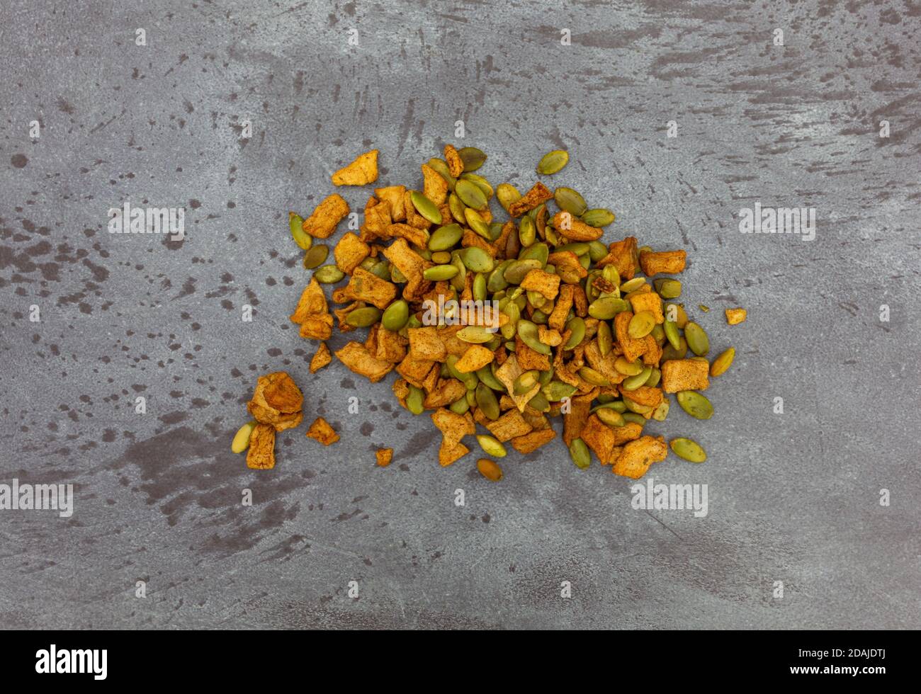 Vista dall'alto di una piccola porzione di semi di zucca torrefatti organici e pezzi di mela disidratati su un piano portapaziente con chiazze grigie. Foto Stock