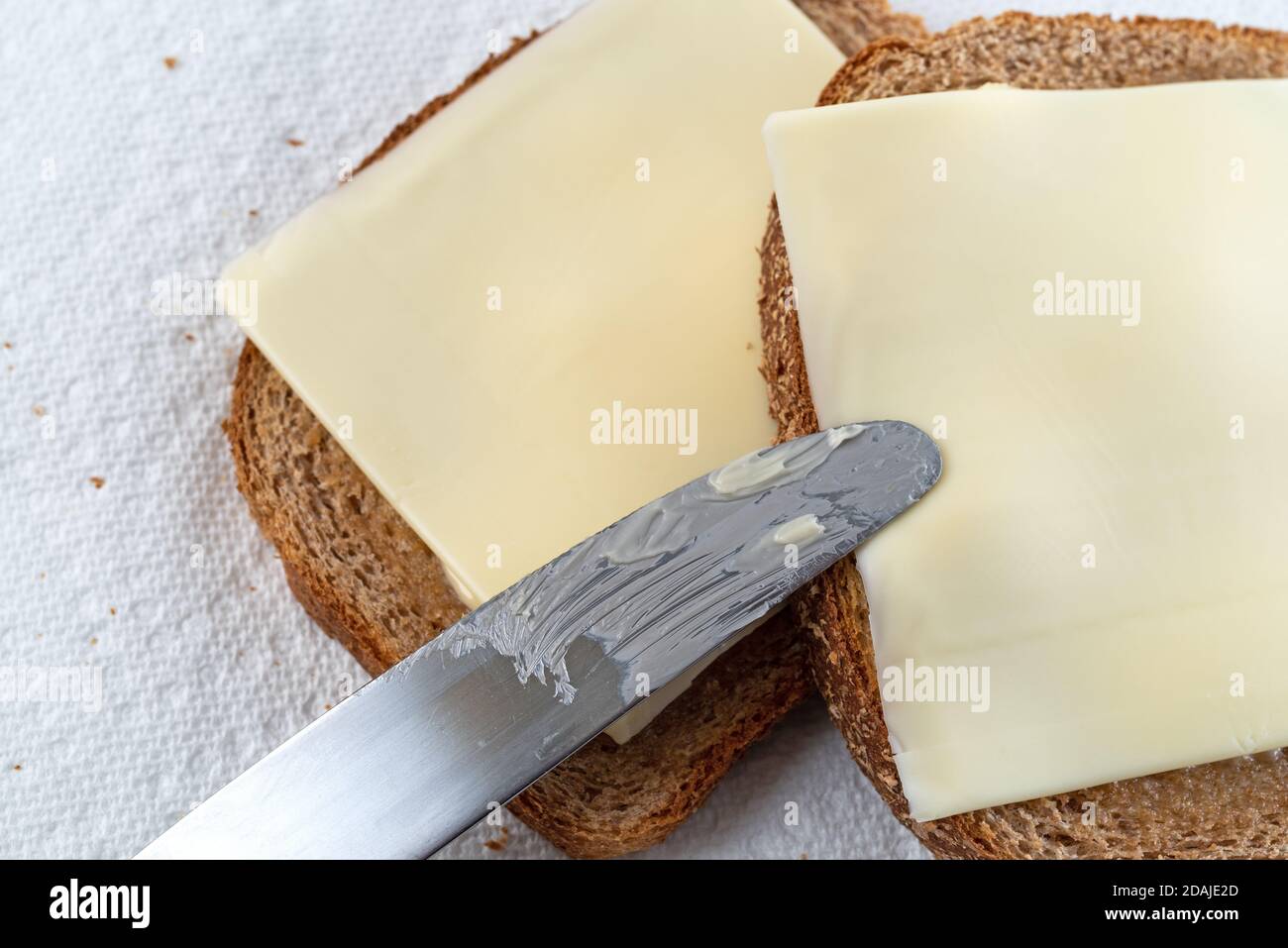 Vista dall'alto di un coltello da burro in cima a due fette di pane di grano con margarina e formaggio su teli di carta illuminati da luce naturale. Foto Stock