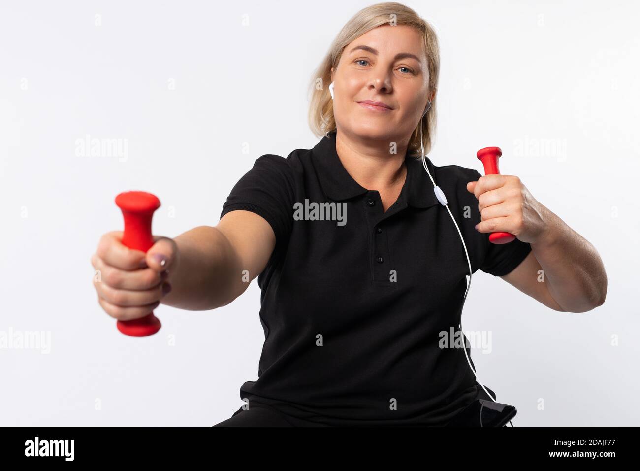 Donna anziana che fa sport a casa con i manubri ascolta la musica con le cuffie. Concetto di fitness durante una pandemia. Foto su sfondo bianco. Foto Stock