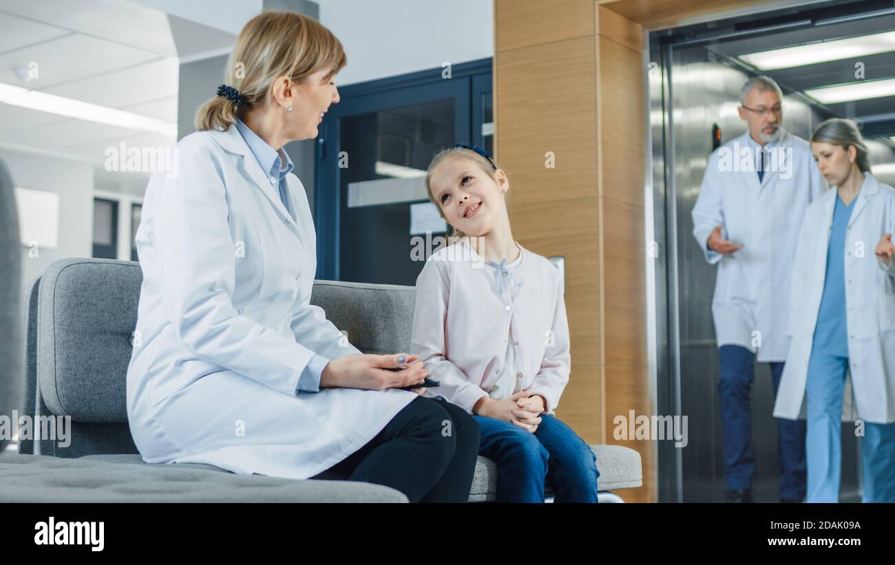 Nella lobby dell'Ospedale Female Doctor parla con una piccola ragazza carina mentre si siede sul divano. Ospedale moderno occupato con la migliore Pediatria possibile Foto Stock